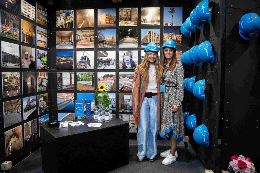dvije djevojke s plavim kacigama stoje u prostoriji u kojoj na zidu visi mnogo plavih kaciga i fotografija
