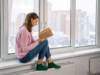 Devojka sedi kod prozora i cita knjigu